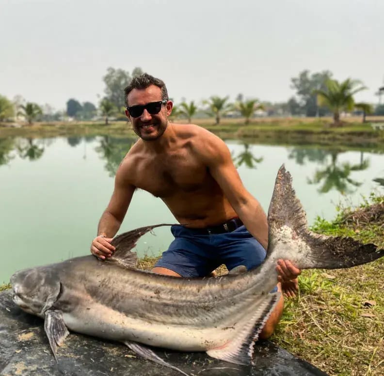 Tom Williams kneeling at the edge of a pond, holding up an enormous catfish.