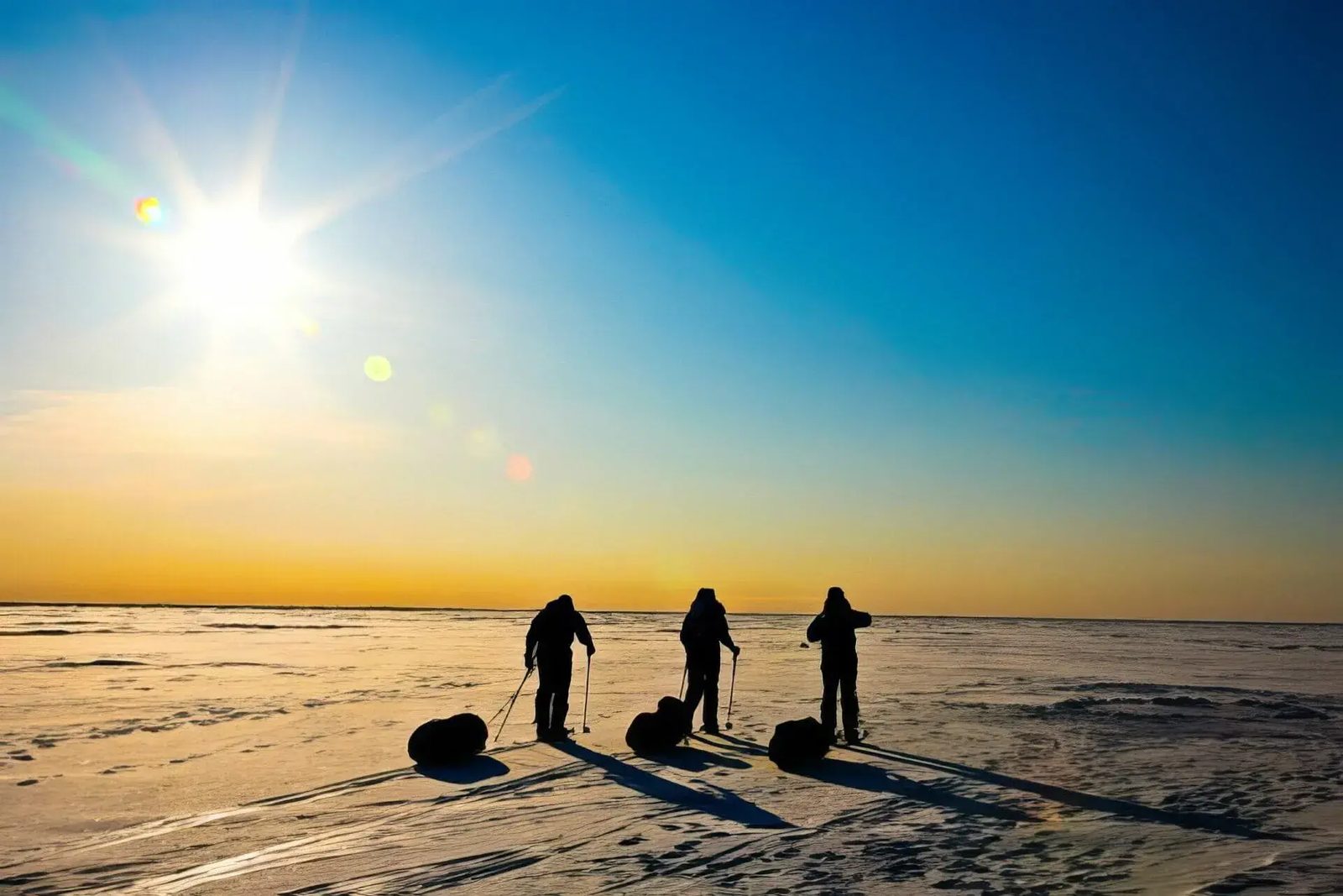 Three expedition members silhouetted against a low sun on a frozen plain, hauling sleds behind them.