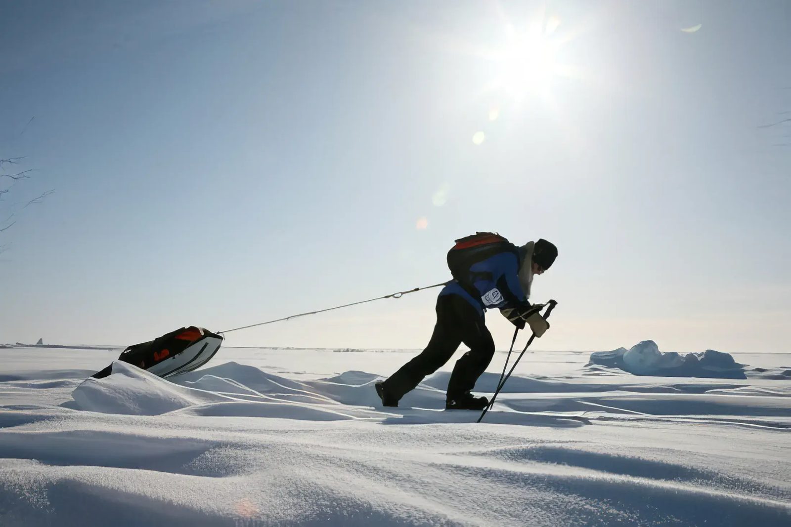 A polar trekker leaning hard into a harness to haul a sled across bright sunlit snow.