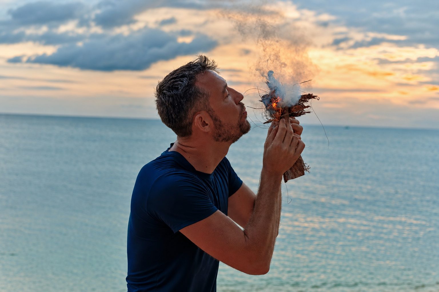 Tom Williams crouches on a beach at sunset, blowing a bundle of tinder into flame with the sea and clouded sky behind him.