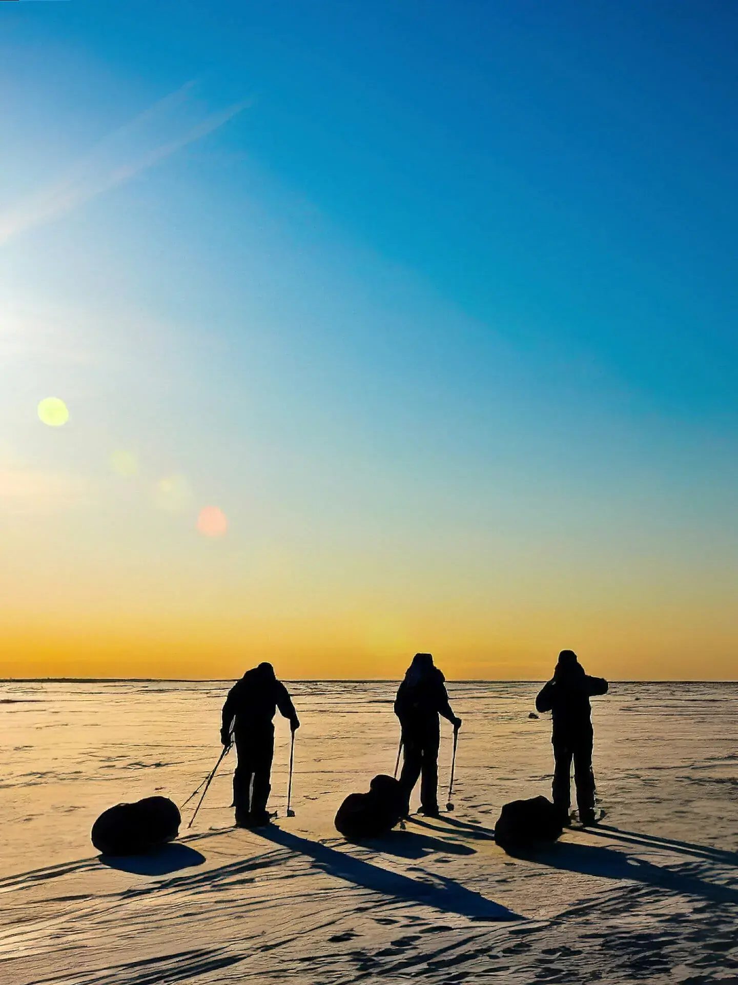 Three figures with sleds silhouetted against a golden sunset on a frozen plain.